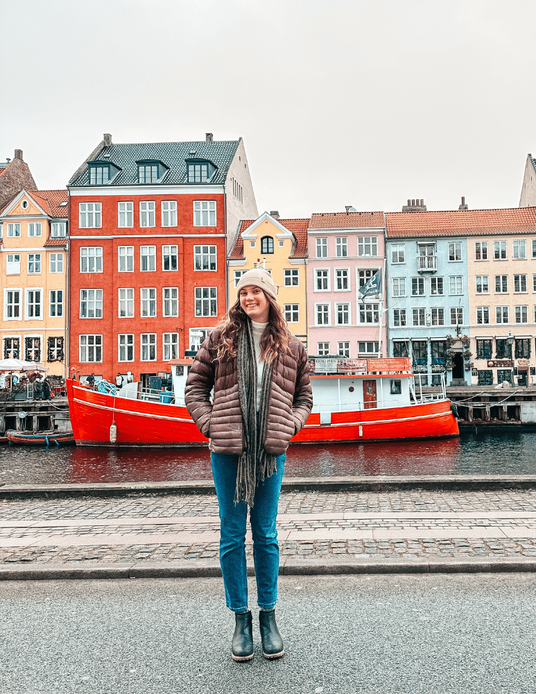Camrie standing in front of the iconic colorful buildings of Nyhavn in Copenhagen, Denmark, wearing a brown puffer jacket and beanie
