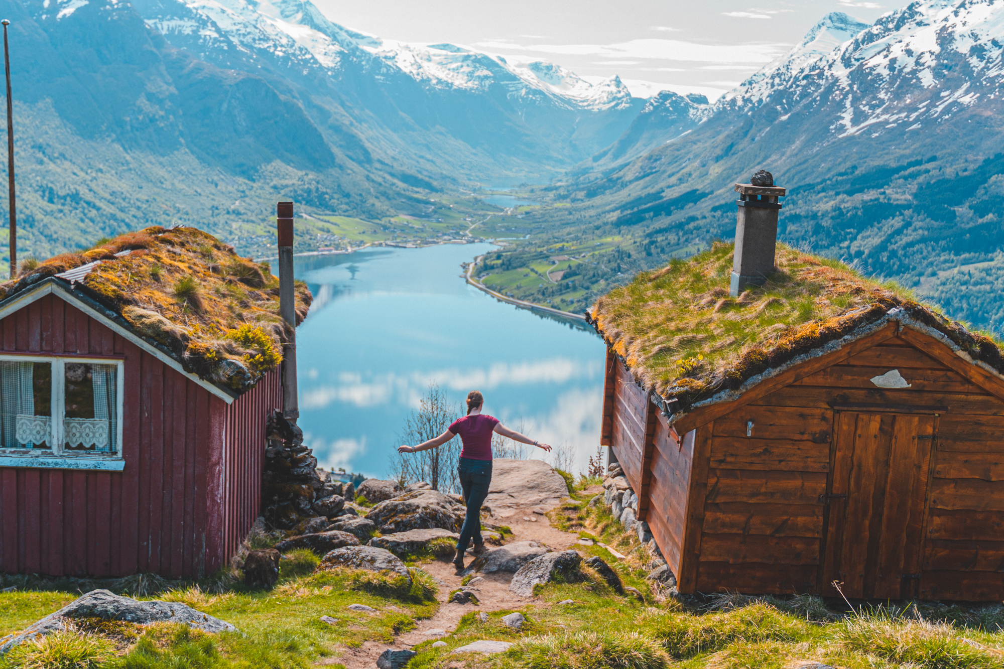Camrie with arms outstretched between traditional Norwegian turf-roof houses overlooking a stunning fjord and snow-capped mountains